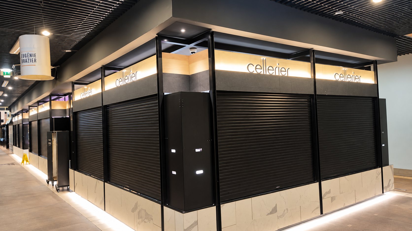 Angle d'une boutique Cellerier dans une galerie couverte, avec rideaux métalliques noirs baissés, enseignes lumineuses et soubassement en marbre clair.