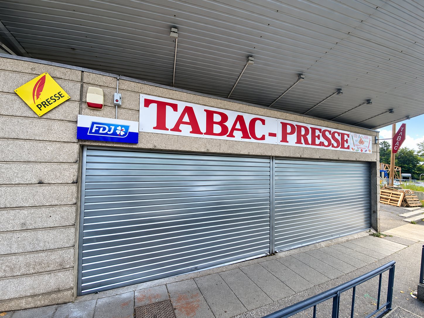 Façade d'un tabac-presse en pierre avec enseigne rouge, logos Presse et FDJ et un rideau métallique baissé.