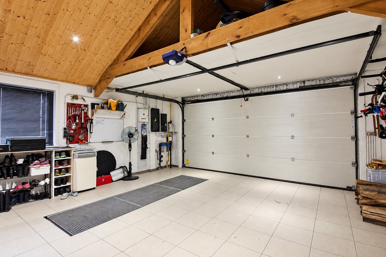 Intérieur d'un garage carrelé avec une grande porte sectionnelle blanche motorisée, une charpente en bois apparente, un établi d'outils et des étagères à chaussures.