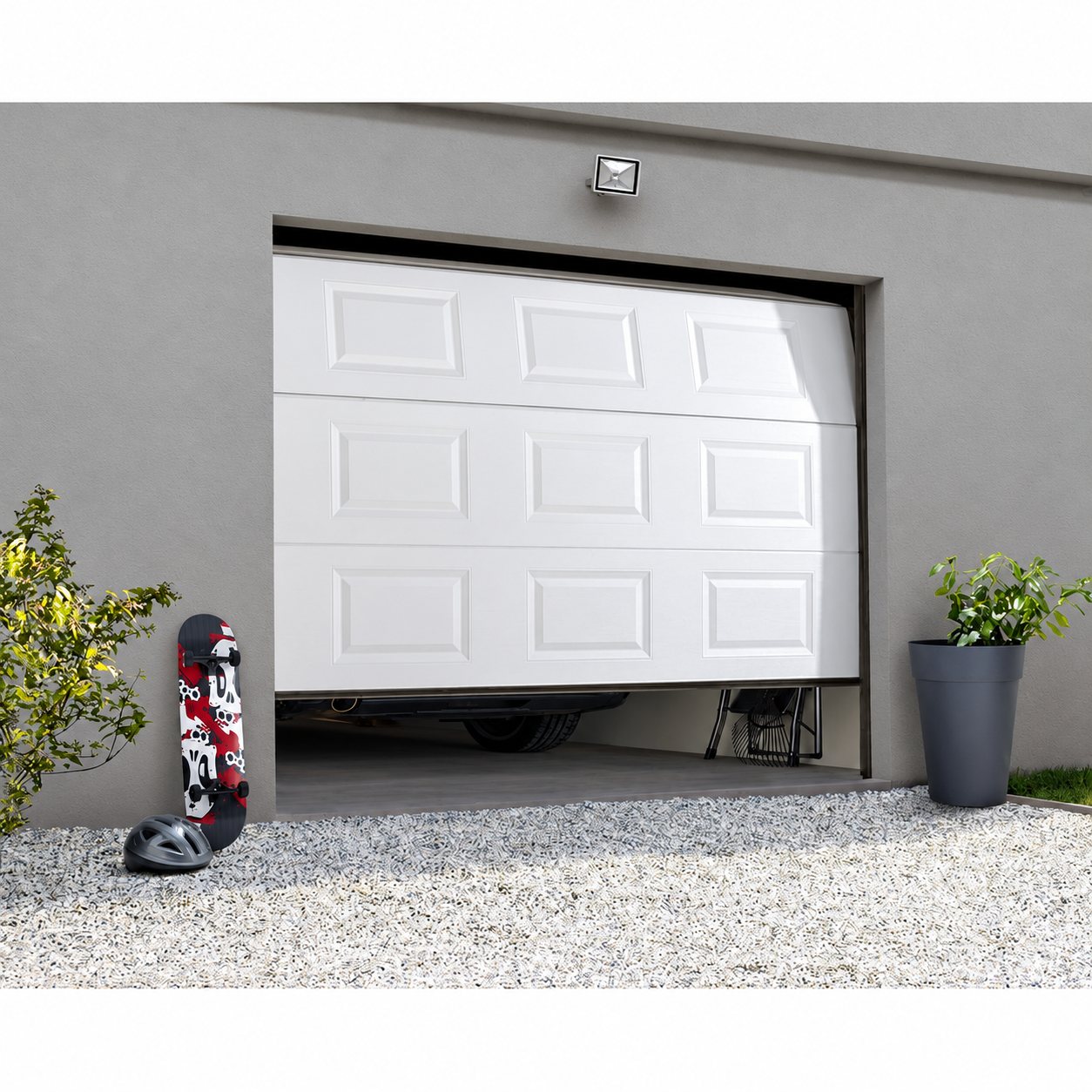 Une porte de garage sectionnelle blanche à panneaux, partiellement relevée, sur une façade grise avec un skateboard, un casque et des plantes devant le gravier.