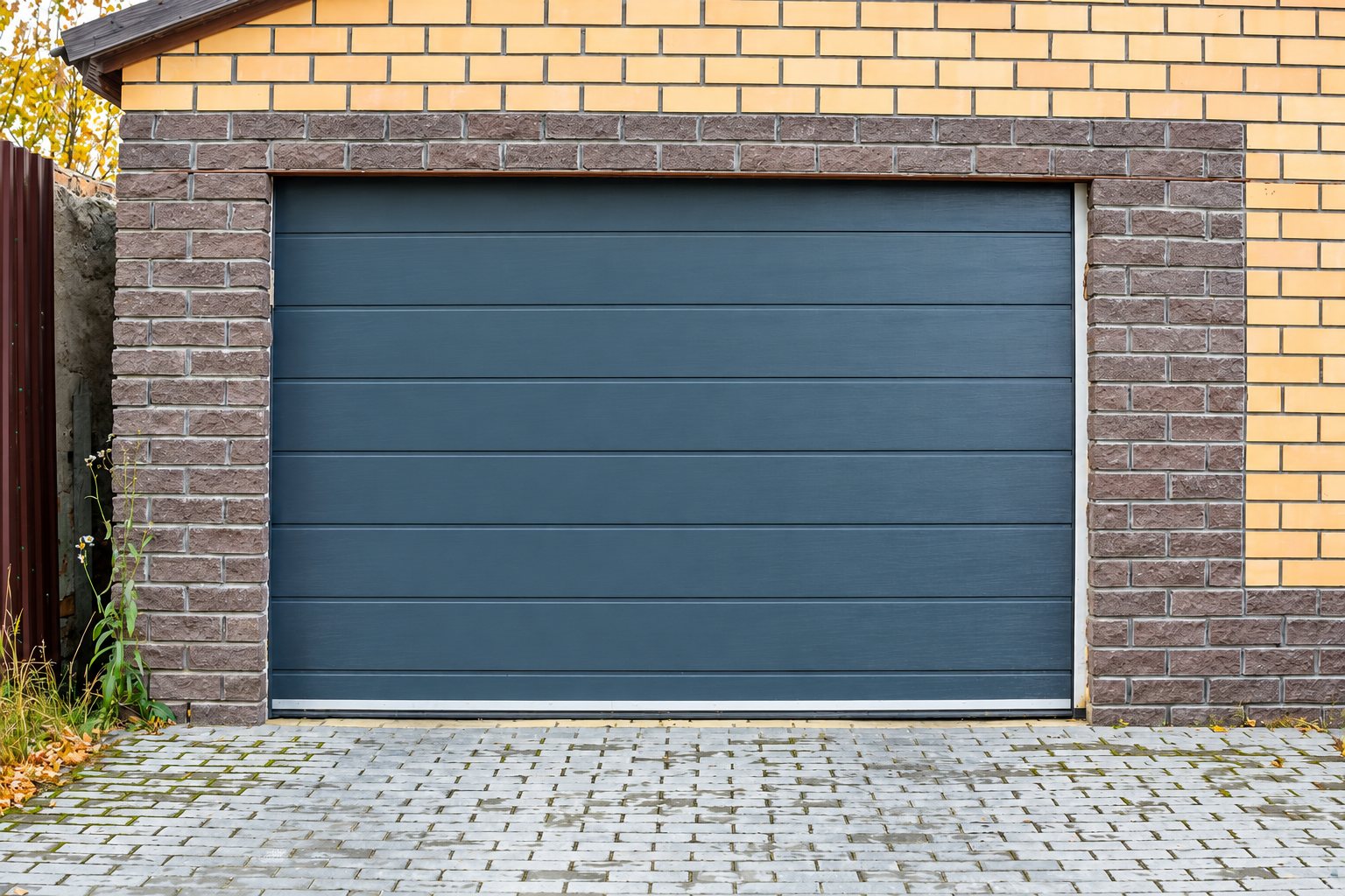 Une porte de garage sectionnelle gris anthracite installée dans un encadrement de briques beiges et brunes, devant une allée pavée.