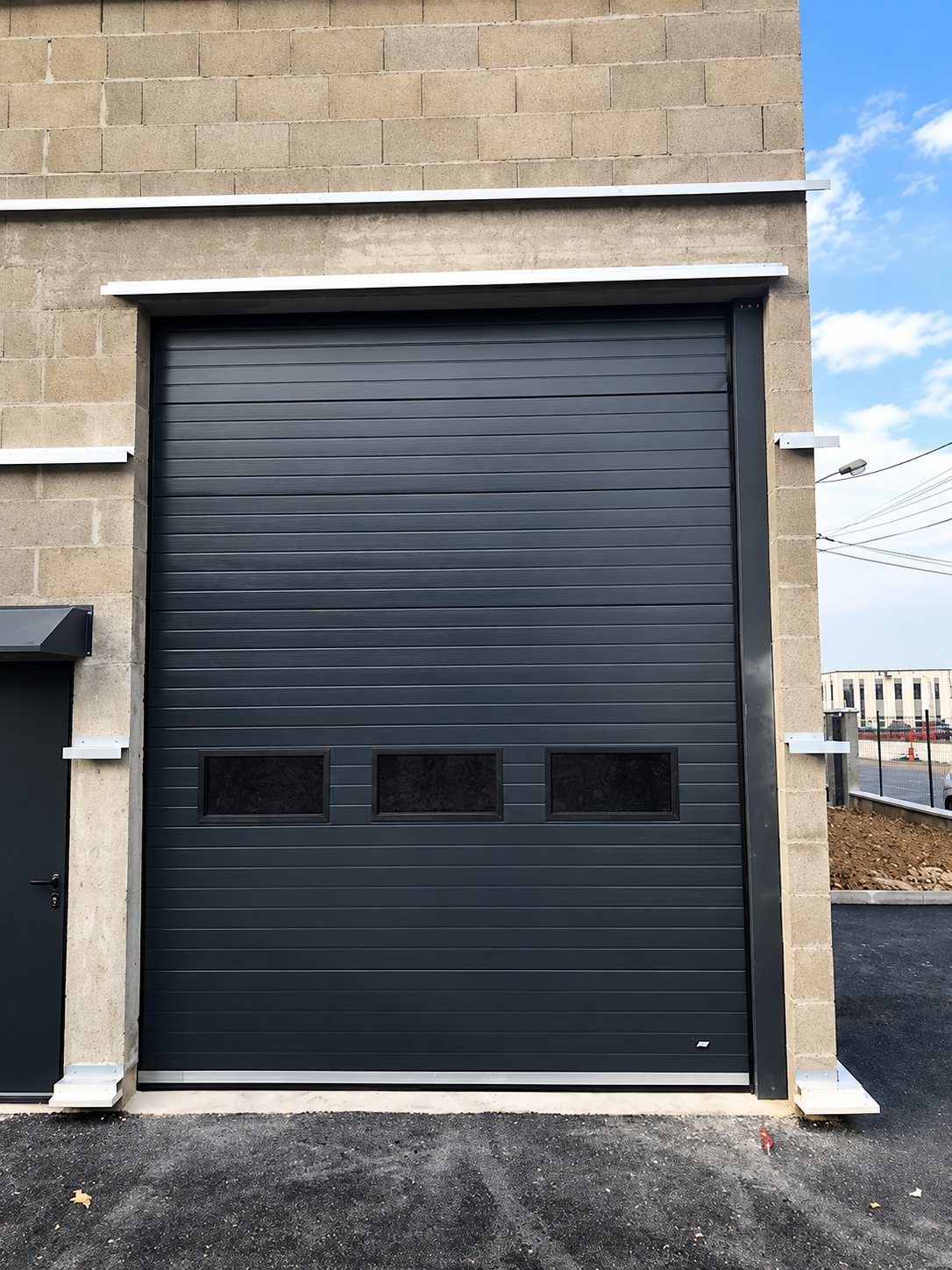 Grande porte sectionnelle anthracite à trois hublots installée dans la façade en parpaings d'un bâtiment neuf, à côté d'une porte de service.