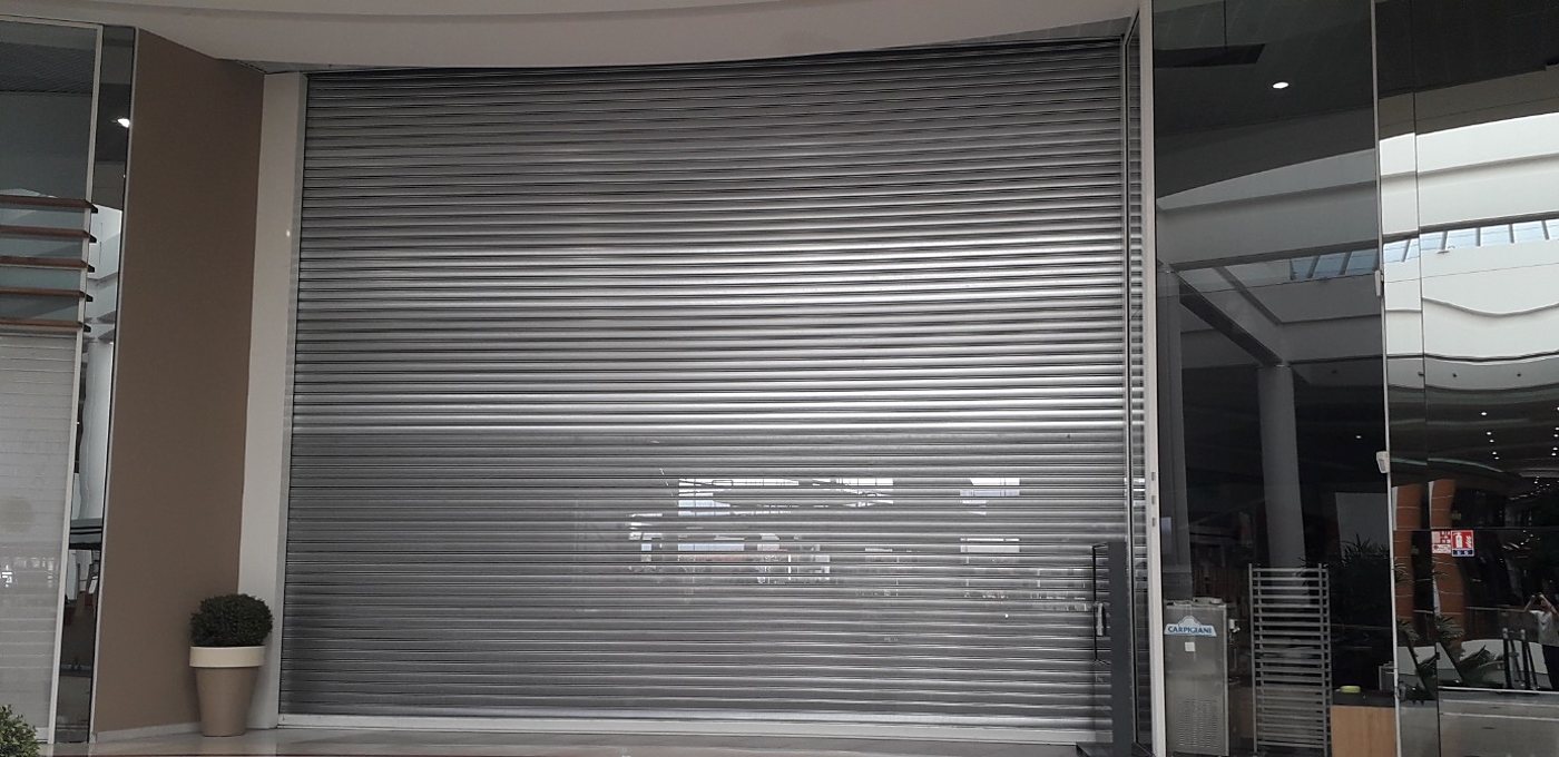 Un grand rideau métallique gris fermé devant une boutique d'un centre commercial, à côté d'une plante en pot et de vitrines en verre.