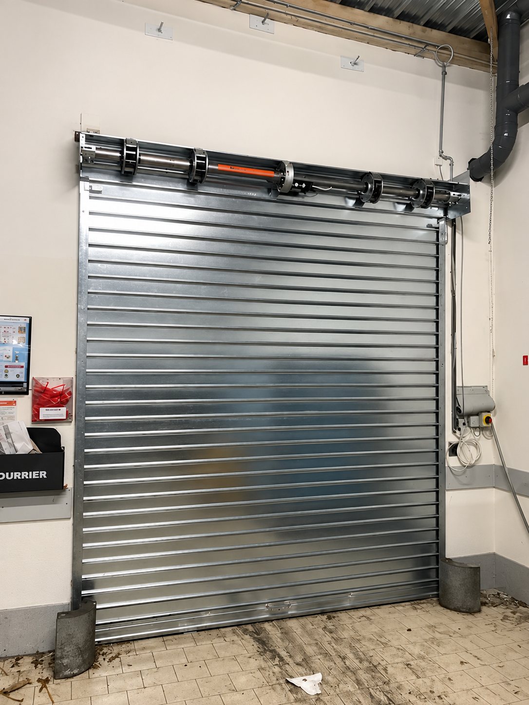Un rideau métallique galvanisé à lames horizontales, équipé de son moteur en partie haute, installé dans un local au sol carrelé et au mur blanc.