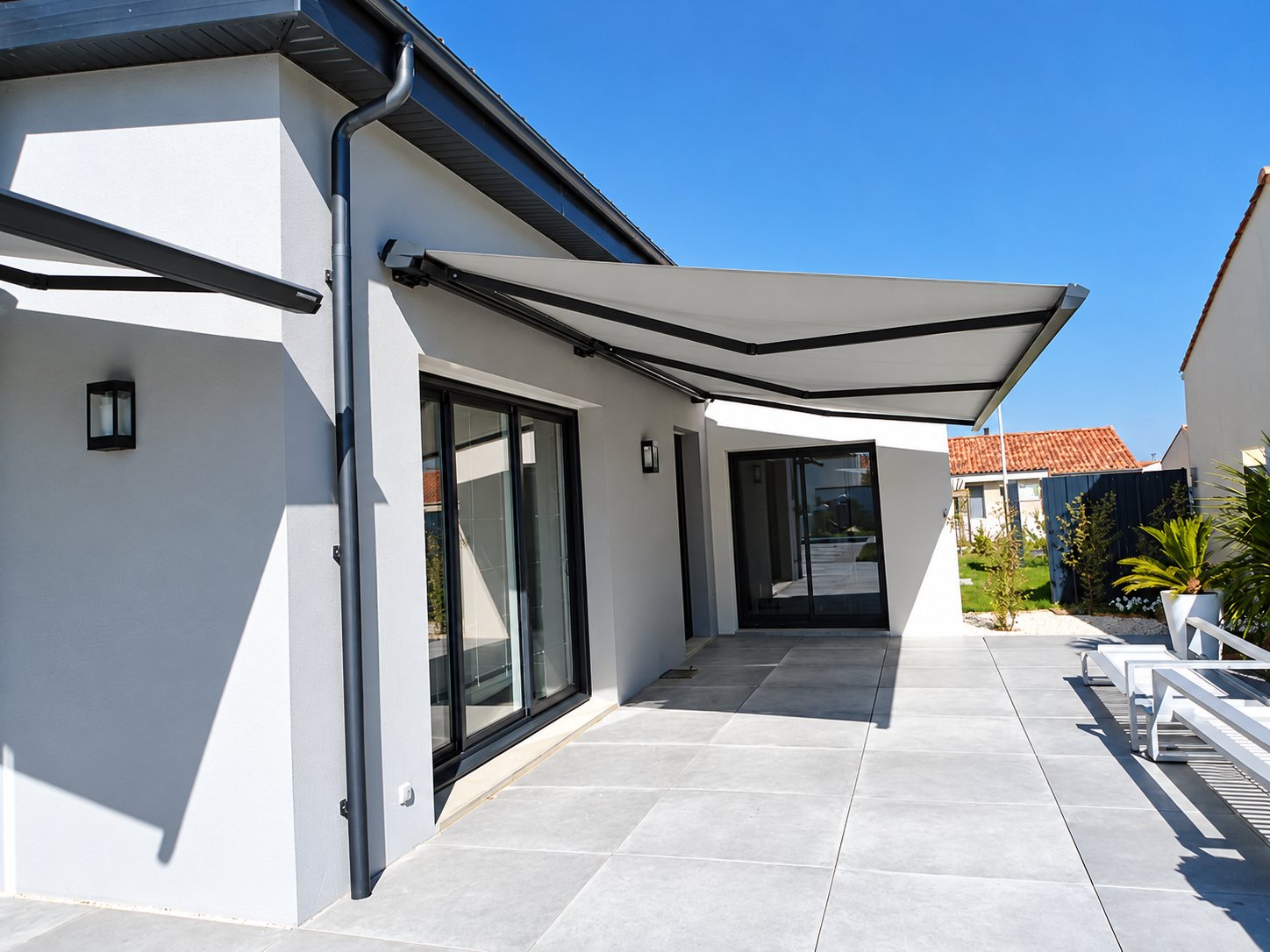 Un store banne gris déployé au-dessus d'une terrasse carrelée devant une maison moderne aux baies vitrées sombres, sous un ciel bleu.