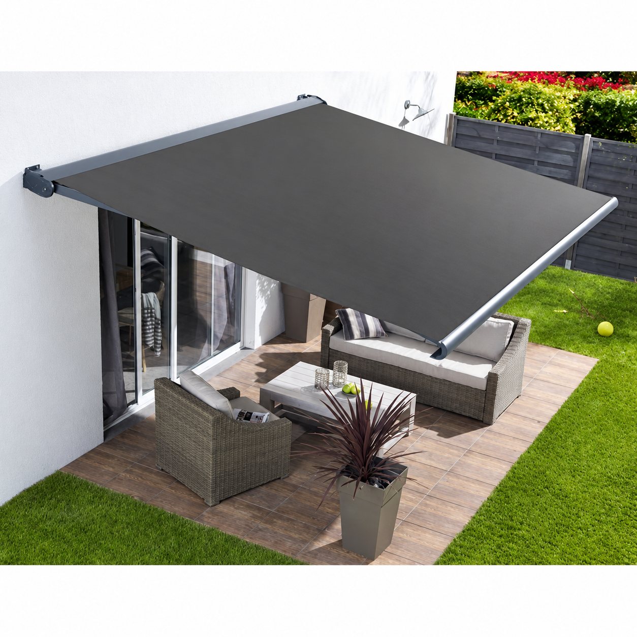 Un store banne gris anthracite déployé sur une terrasse en dalles où sont installés un canapé et un fauteuil en résine tressée, une table basse et une plante en pot.